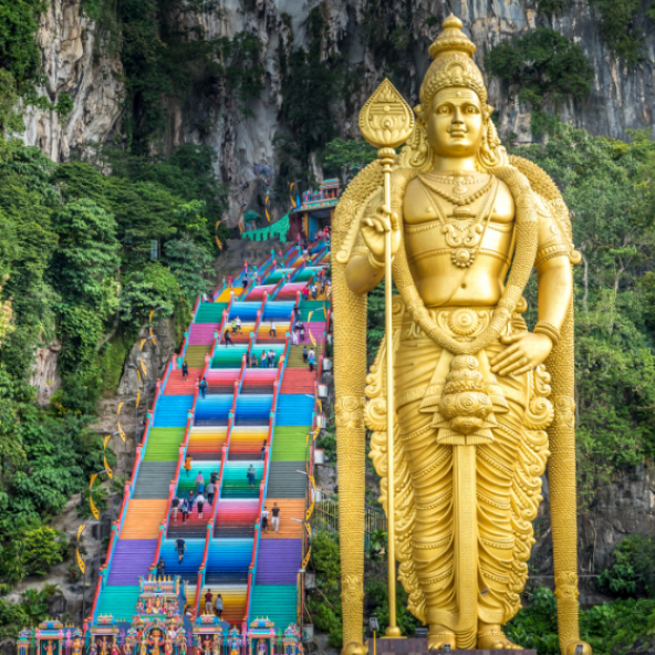 A large golden standing statue of a deity with an ornate robe, holding a staff, in front of a steep cliffside; a long colorful staircase with many small figures and railings runs behind it.