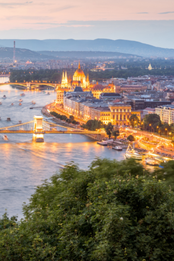 Wide aerial view of a river at sunset, with a long bridge, calm water, and illuminated buildings including a golden cathedral and tall tower in the distance.