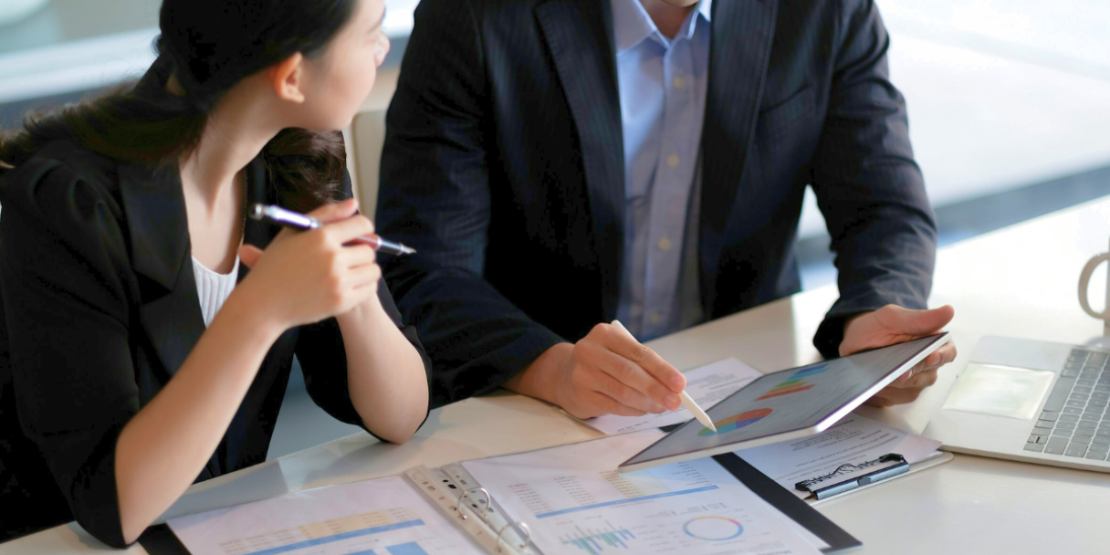 two individuals in business attire reviewing charts and graphs on a tablet and papers spread across a desk