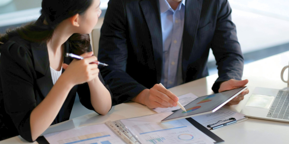 two individuals in business attire reviewing charts and graphs on a tablet and papers spread across a desk