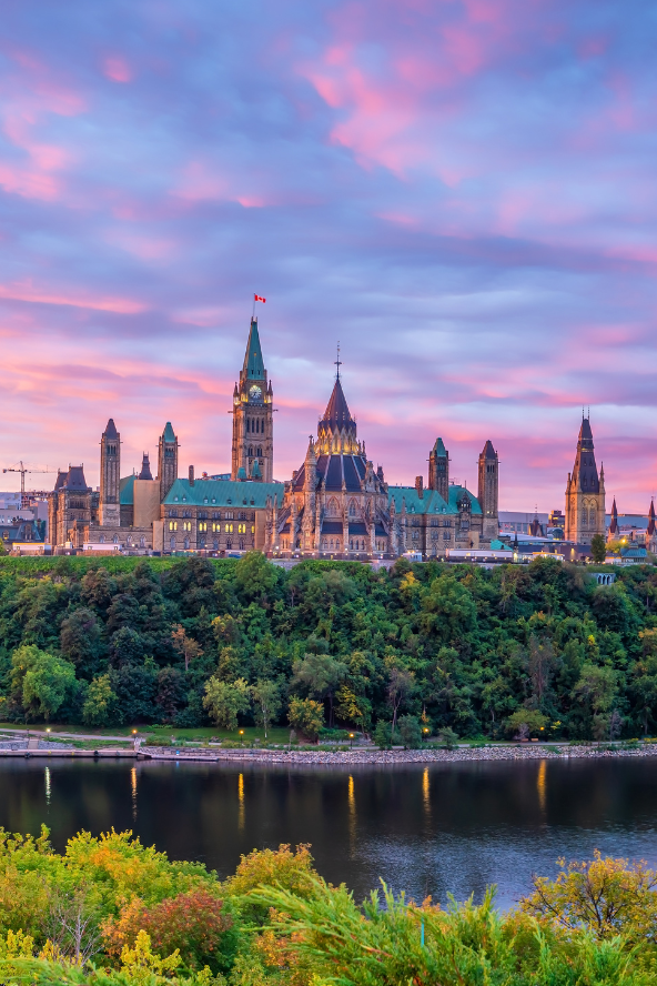 the Parliament Hill buildings in Ottawa, Canada, are silhouetted against a vibrant pink and purple sunset sky, with a river and lush green trees in the foreground