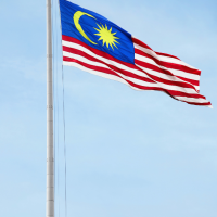 A tall flagpole flying a Malaysian flag with a blue field, yellow crescent and star, and red-and-white wavy stripes against a clear blue sky.