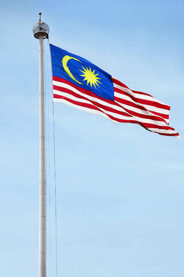 A tall flagpole flying a Malaysian flag with a blue field, yellow crescent and star, and red-and-white wavy stripes against a clear blue sky.