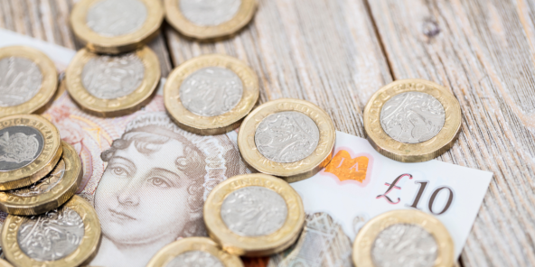 Gold-edged £1 coins scattered on a wooden table beside a UK ten-pound note showing a £10 value.