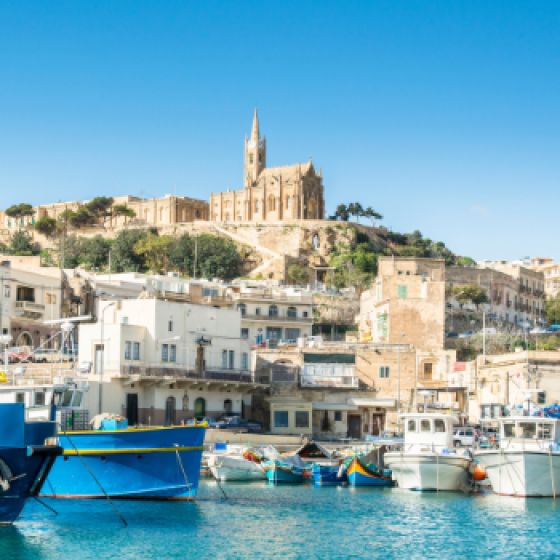 Harbour water in the foreground with moored boats and a blue docked vessel, overlooking a white seaside town on a hill with a prominent church tower under a clear blue sky.