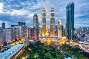 A twilight city skyline with three illuminated golden skyscraper towers in the centre, surrounding high-rise buildings, a river and green park below, and a blue cloudy sky.