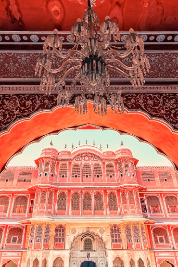Ornate chandelier hanging in a richly decorated hall above a large arched view of a historic building lit with red light, with patterned walls and decorative borders framing the scene.