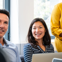 Three people are gathered around laptops in a bright room. A person in a blue shirt looks to the left, a person in a striped shirt smiles towards the camera, and a person in a yellow dress holds a folder.