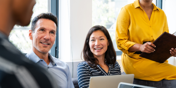 Three people are gathered around laptops in a bright room. A person in a blue shirt looks to the left, a person in a striped shirt smiles towards the camera, and a person in a yellow dress holds a folder.