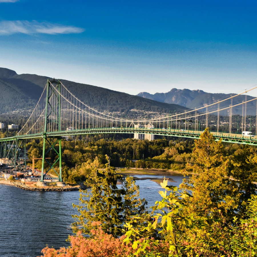 a suspension bridge with green towers and cables spans across a body of water, with forested hills and mountains in the background under a bright blue sky