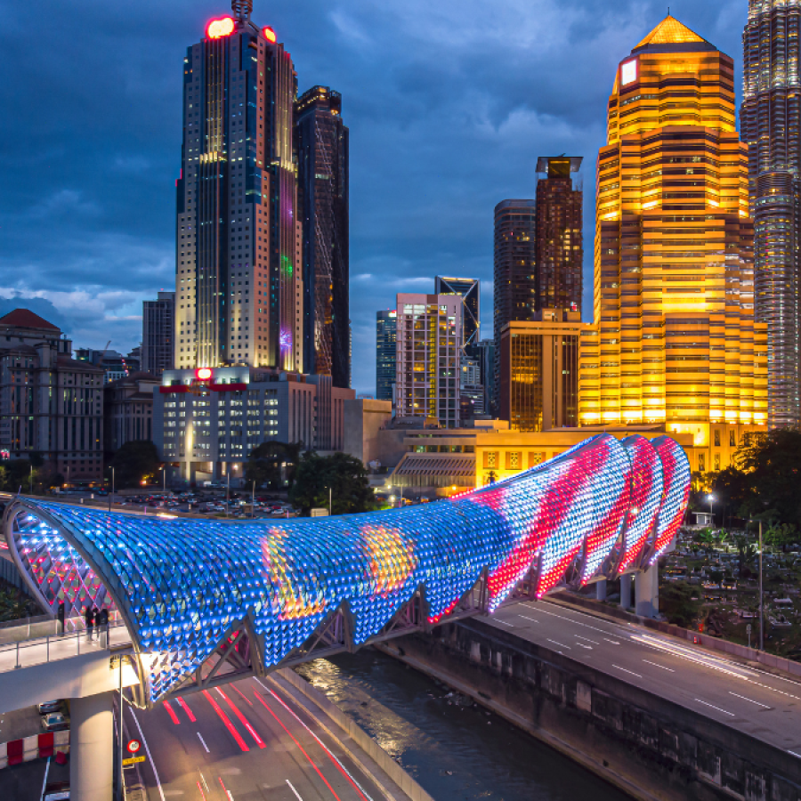 Night skyline with tall skyscrapers and a brightly lit curved bridge covered in blue and red lights, with traffic moving along the road beneath under a dark blue sky.
