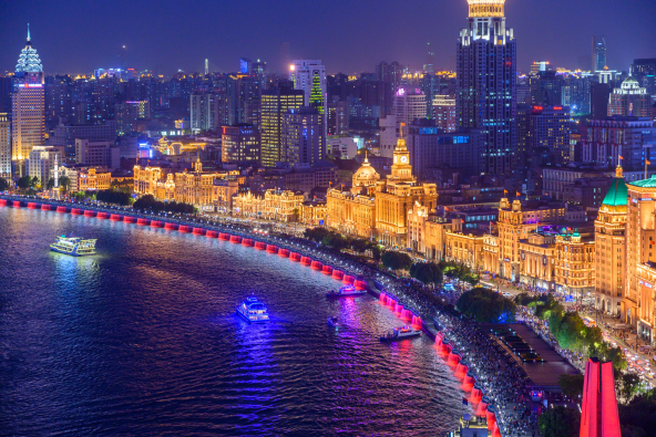 ighttime skyline along a wide river, with illuminated high-rise buildings and a lit promenade lined by red-and-white lights, under a deep blue sky.