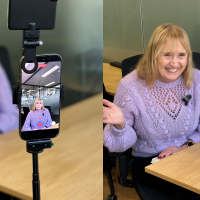 Mauve CEO Ann in a purple sweater sits at a desk, waving at a phone mounted on a tripod with a small light attached, which is recording her