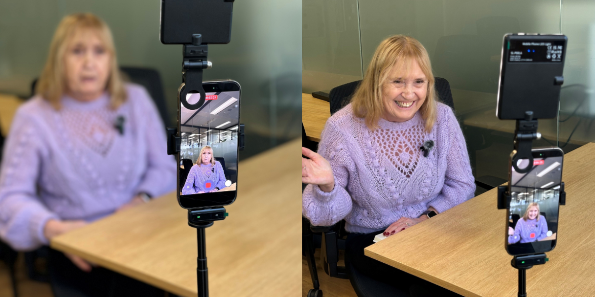 Mauve CEO Ann in a purple sweater sits at a desk, waving at a phone mounted on a tripod with a small light attached, which is recording her