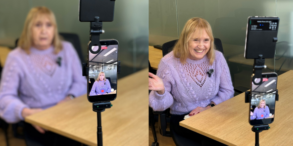 Mauve CEO Ann in a purple sweater sits at a desk, waving at a phone mounted on a tripod with a small light attached, which is recording her