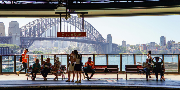 People wait on a platform at Circular Quay, with Sydney Harbour Bridge and city skyline in the background.