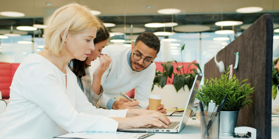 three people gathered around a laptop at a desk in an office, with one person typing and the other two looking at the screen