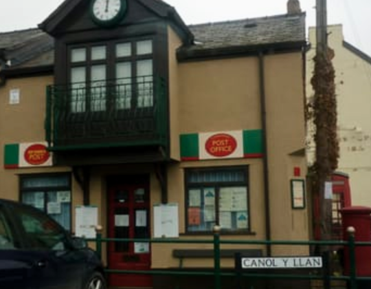 a post office building with a clock above the entrance, Post Office signs, a balcony, and a red telephone box nearby