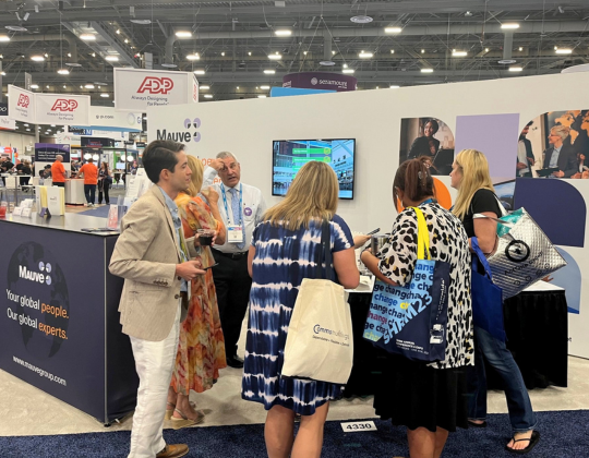 A group of people gathered around a trade show booth with "Mauve" branding, featuring images and a screen displaying information.