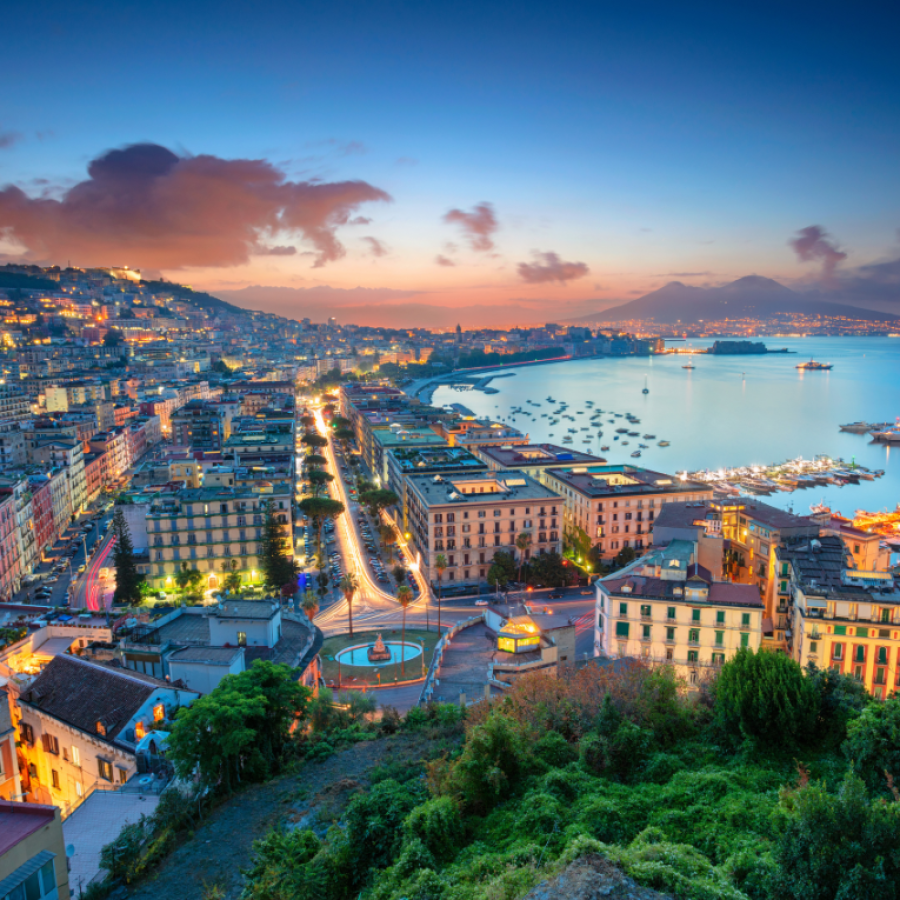 Panoramic coastal city view at dusk with warm street lights, terracotta rooftops, a curving waterfront promenade, and a calm blue harbour dotted with boats and marina piers.