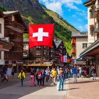 a bustling street scene in a European mountain town, with traditional wooden buildings, shops, and a large Swiss flag hanging overhead. Many people are walking along the street under a bright blue sky with some clouds