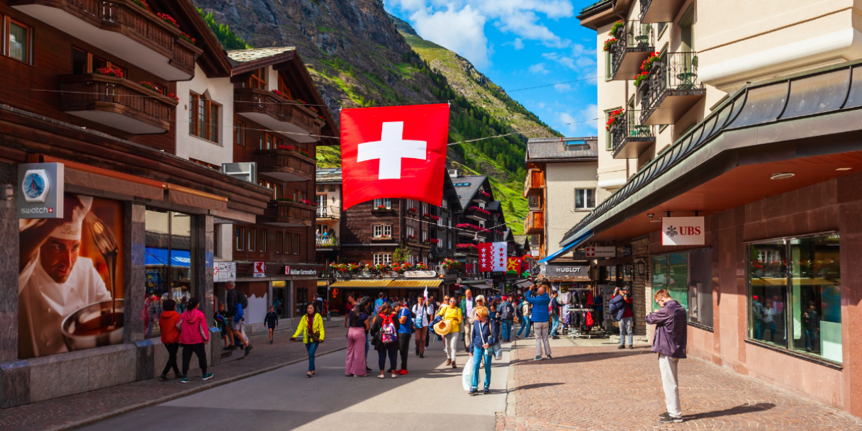a bustling street scene in a European mountain town, with traditional wooden buildings, shops, and a large Swiss flag hanging overhead. Many people are walking along the street under a bright blue sky with some clouds