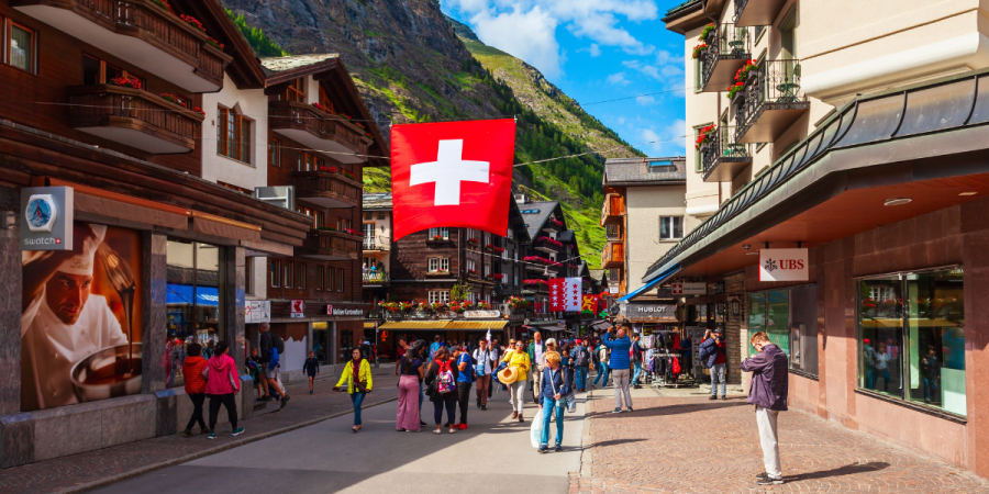 a bustling street scene in a European mountain town, with traditional wooden buildings, shops, and a large Swiss flag hanging overhead. Many people are walking along the street under a bright blue sky with some clouds