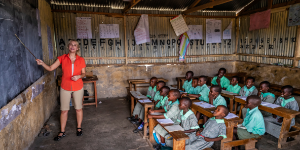 Woman in a red shirt stands beside a classroom with multiple children seated at wooden desks, facing forward, inside a rustic room with handwritten wall posters and a corrugated metal ceiling.