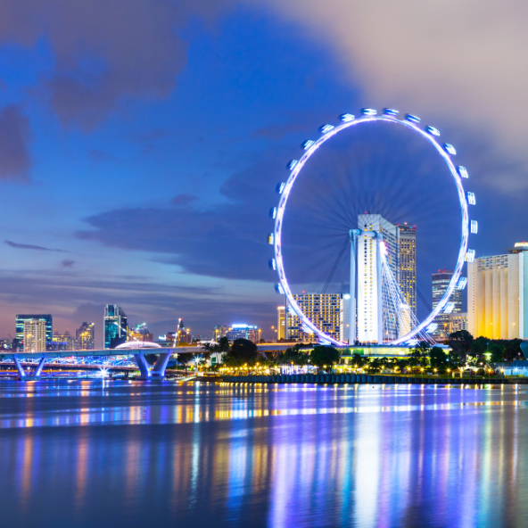 a cityscape at dusk with a large Ferris wheel illuminated by white lights, reflecting on the water in the foreground