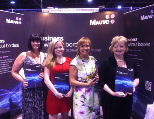 four women stand in front of a booth with "Mauve" branding and "business without borders" text. They each hold a brochure with a blue abstract design
