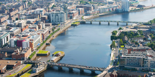 an aerial daytime view of a wide river flowing through a city with bridges, buildings, and greenery along its banks.