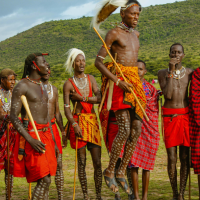 Maasai men in the Masai Mara perform a jumping dance, with one man mid-air wearing a headdress and holding a stick.