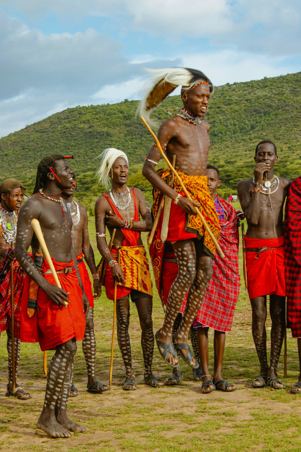 Maasai men in the Masai Mara perform a jumping dance, with one man mid-air wearing a headdress and holding a stick.