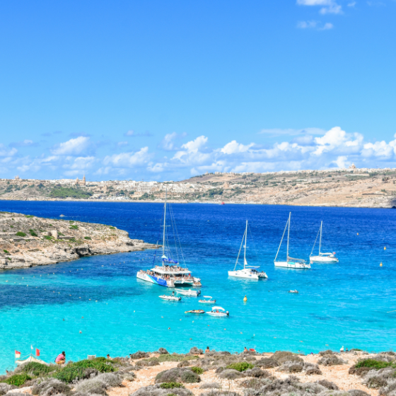 Turquoise cove with a deep-blue harbour, rocky cliffs, and several sailboats and small yachts anchored on calm water under a bright blue sky with scattered clouds.