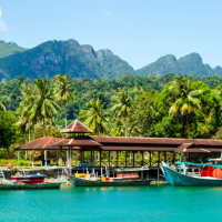 A row of colourful boats moored along a calm turquoise bay beside a wooden dock with a shaded pavilion, with lush palm trees and green mountains rising in the background under a clear blue sky.