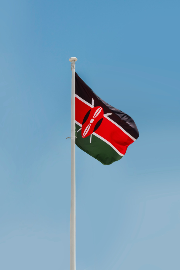 The Kenyan flag waves against a clear blue sky, attached to a white flagpole.