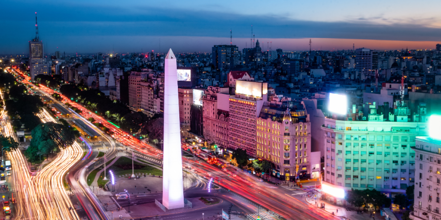 An aerial view of the Obelisco de Buenos Aires at twilight, with light trails from traffic illuminating the wide avenues and surrounding buildings.