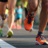 Close-up of runners’ legs sprinting on a road race, with one orange running shoe stepping forward on the asphalt and other participants blurred in the background near traffic cones.