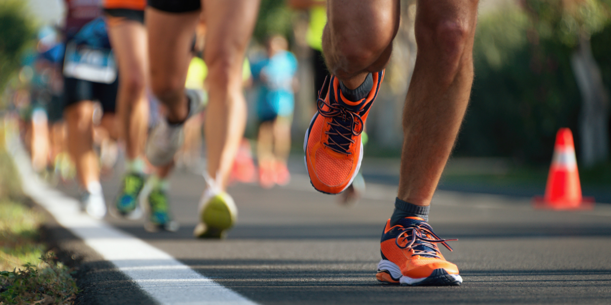 Close-up of runners’ legs sprinting on a road race, with one orange running shoe stepping forward on the asphalt and other participants blurred in the background near traffic cones.