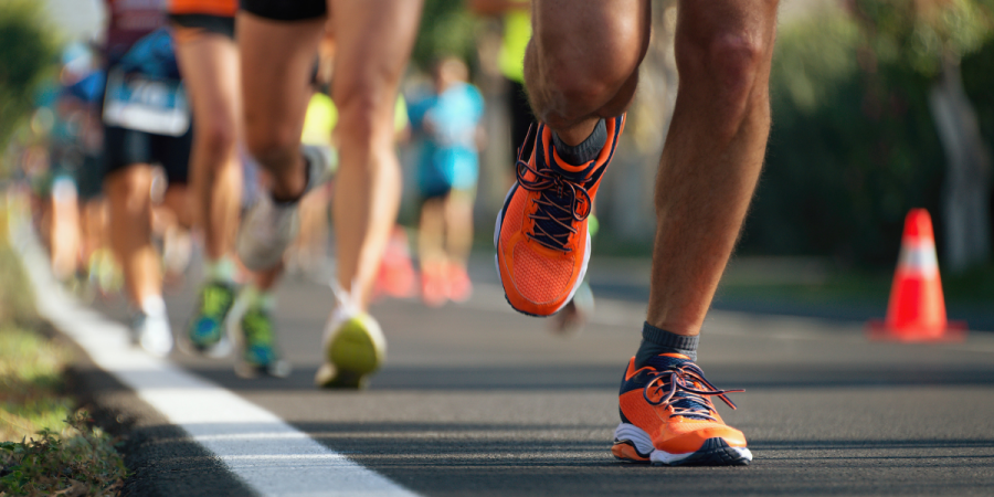 Close-up of runners’ legs sprinting on a road race, with one orange running shoe stepping forward on the asphalt and other participants blurred in the background near traffic cones.