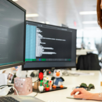 A woman with red hair works at a desk in an office, looking at a computer monitor showing code while two additional screens and office supplies sit nearby.
