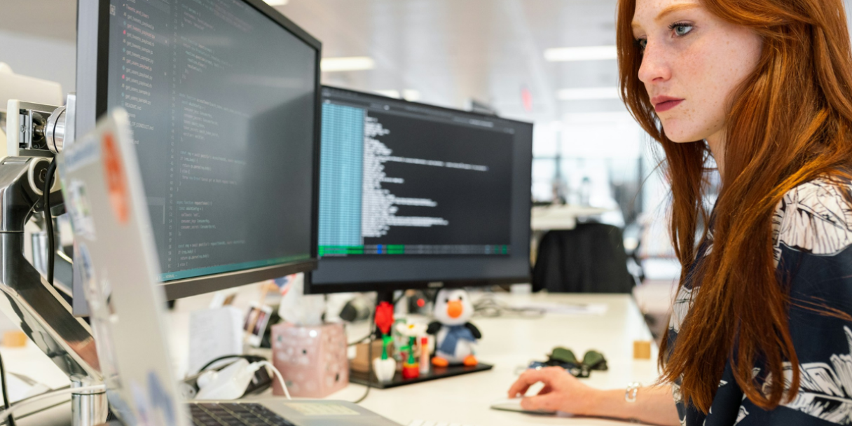 A woman with red hair works at a desk in an office, looking at a computer monitor showing code while two additional screens and office supplies sit nearby.