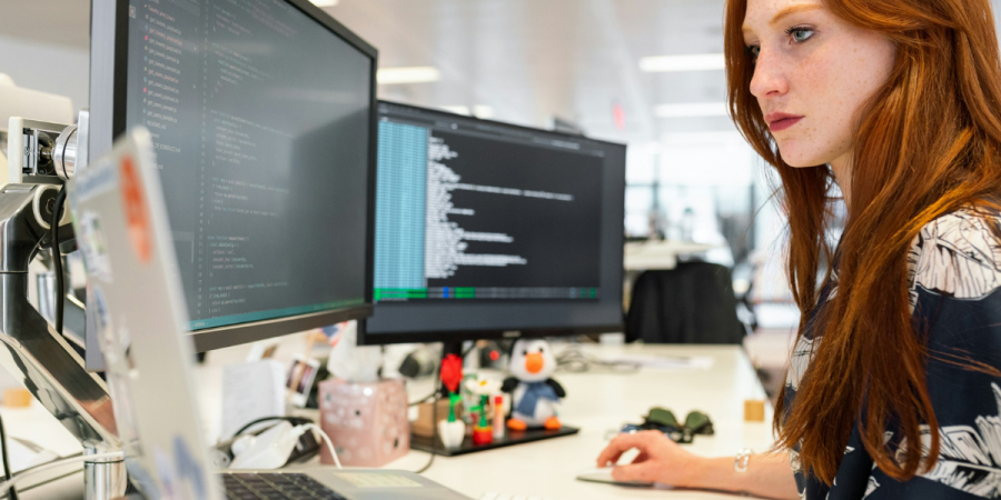 A woman with red hair works at a desk in an office, looking at a computer monitor showing code while two additional screens and office supplies sit nearby.