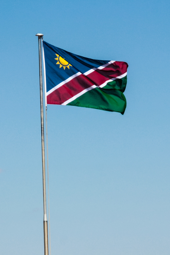 A tall flagpole flying the Namibian flag with green, yellow, black, white, and red colours against a clear blue sky over dry grass and rocky hills.