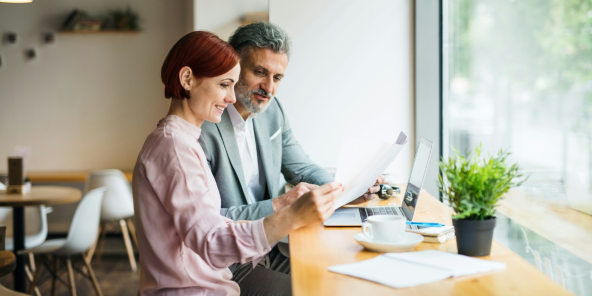 a man and a woman sit at a table looking at papers, with a laptop and coffee cup nearby.