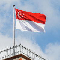 the flag of Singapore, red with a white crescent moon and five white stars, flies against a blue sky with white clouds, above a building with a red tiled roof and decorative metal railing