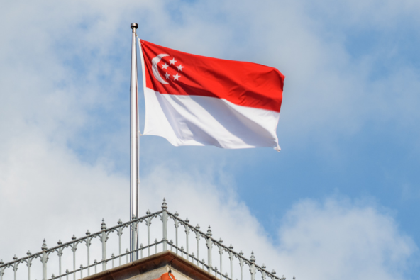 the flag of Singapore, red with a white crescent moon and five white stars, flies against a blue sky with white clouds, above a building with a red tiled roof and decorative metal railing