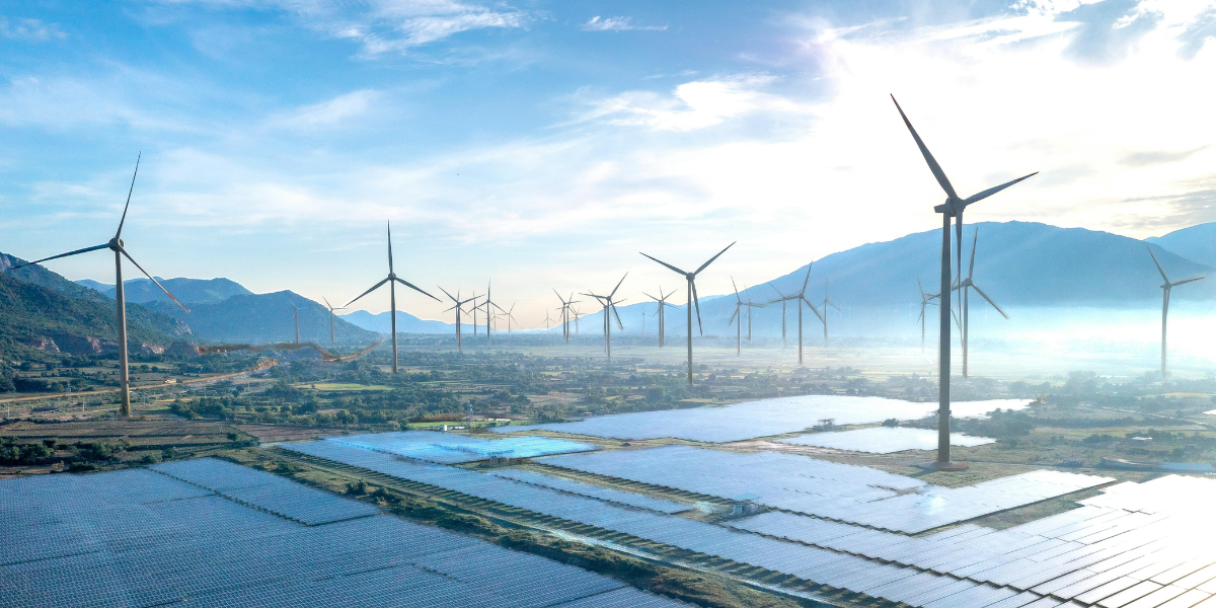 an aerial shot of a landscape with fields of solar panels in the foreground and a row of wind turbines in the background, under a bright, hazy sky