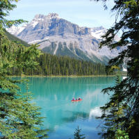 a bright red canoe with two people navigates across a vibrant turquoise lake, framed by lush green pine trees and a majestic, snow-capped mountain range in the background