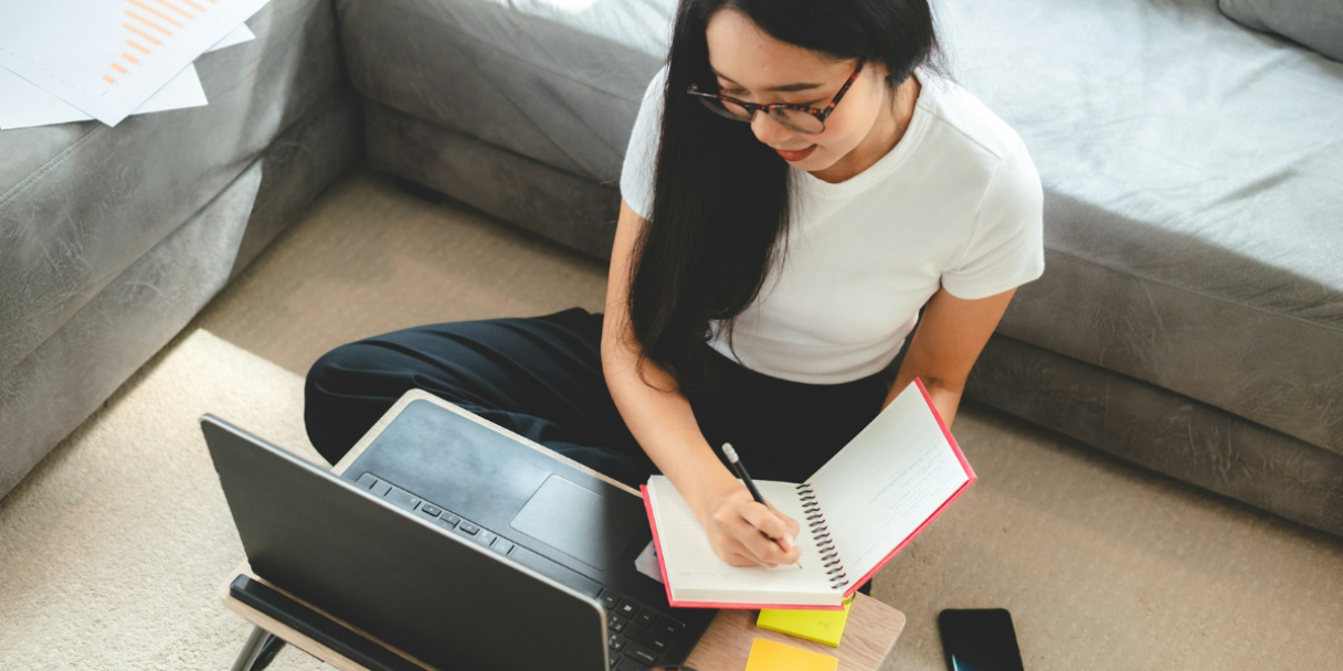 A female employee sitting on the floor with a laptop and notebook, writing in the notebook